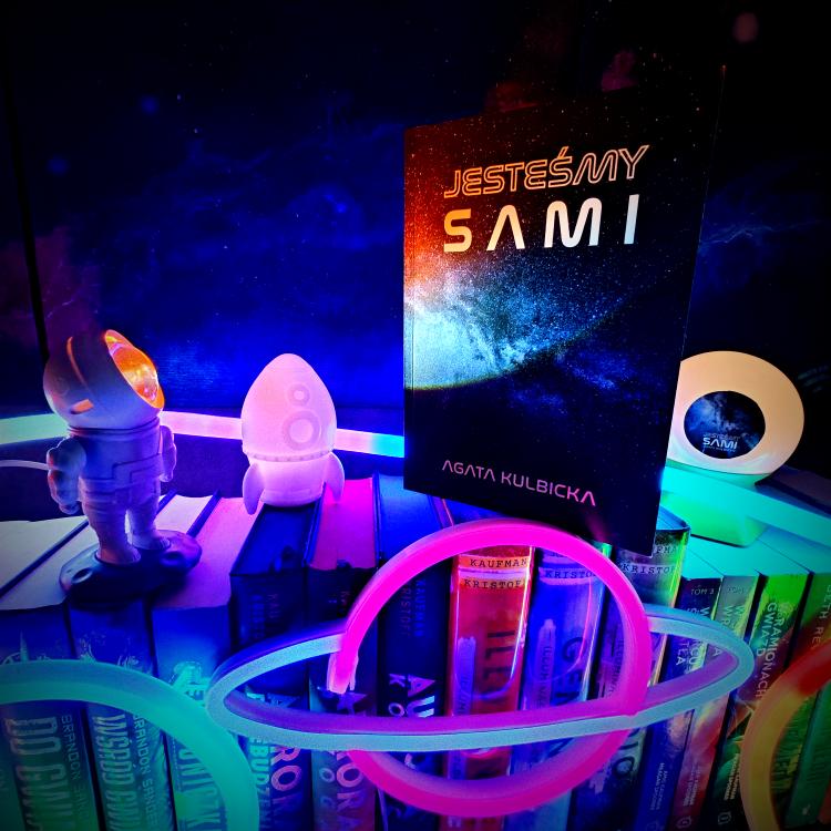 Książka "Jesteśmy sami" w otoczeniu kosmicznych gadżetów: astronauta, rakieta, neon.